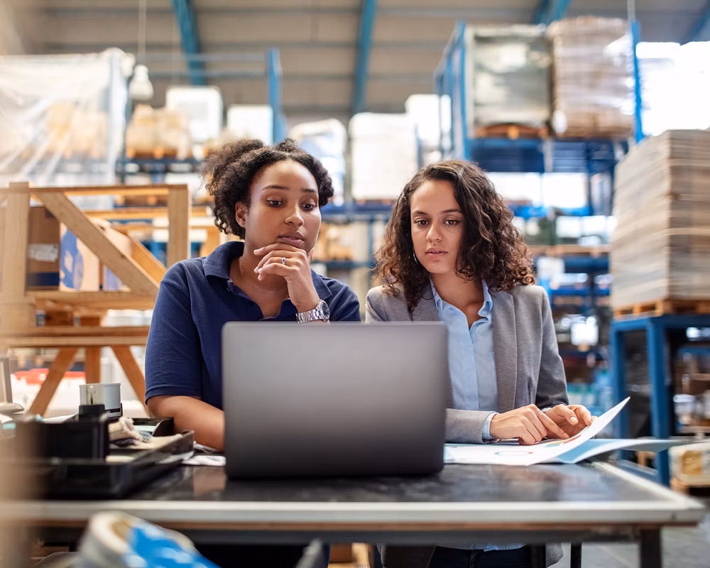 Two young women in a warehouse working on a laptop computer.