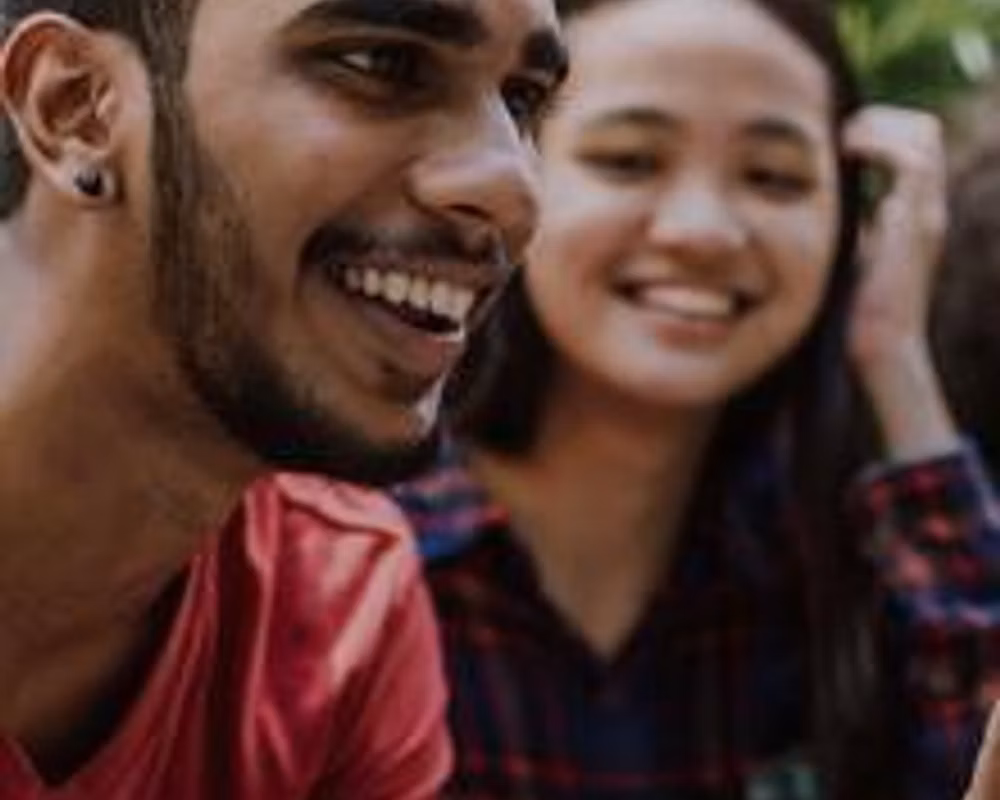 A brown-skinned person with a beard and an East Asian person with long hair sit next to each other smiling