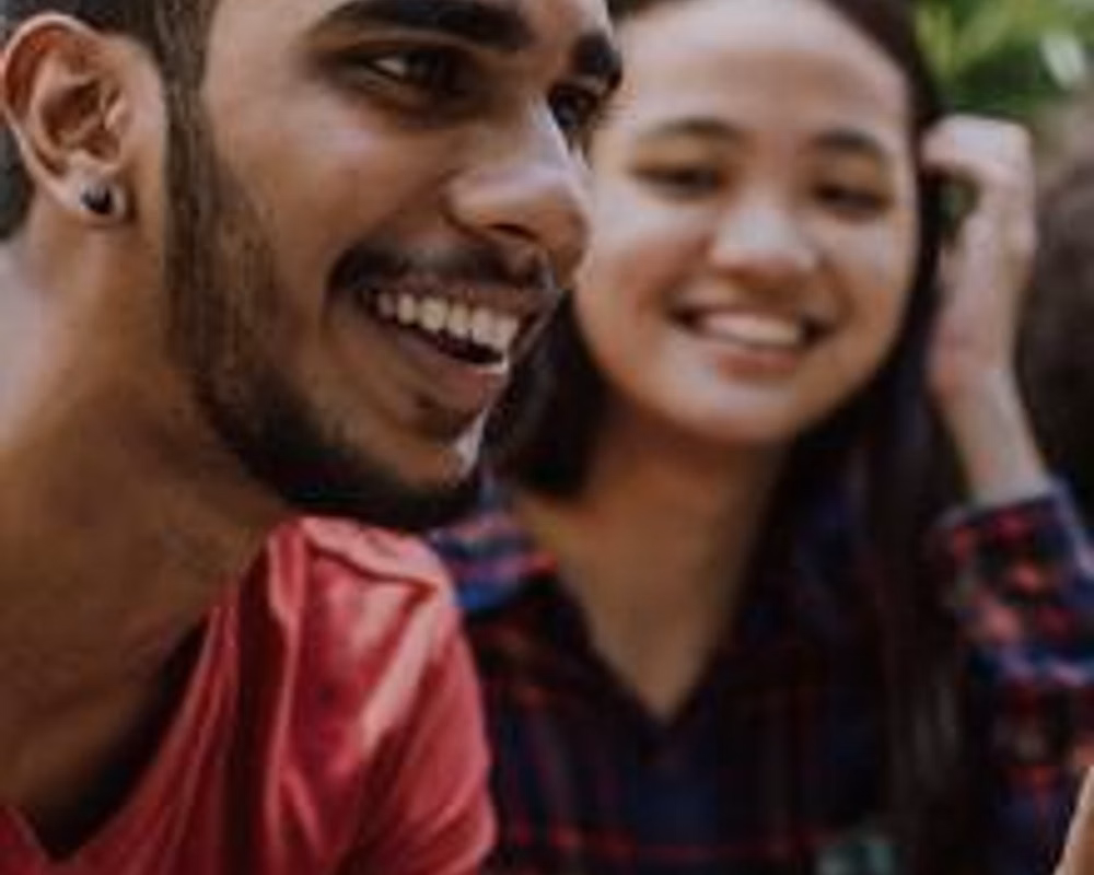 A brown-skinned person with a beard and an East Asian person with long hair sit next to each other smiling