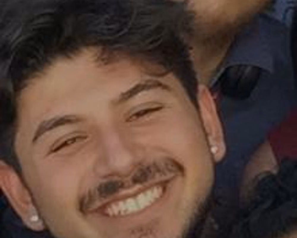 A smiling young person with curly hair, a mustache, a short beard, and earrings