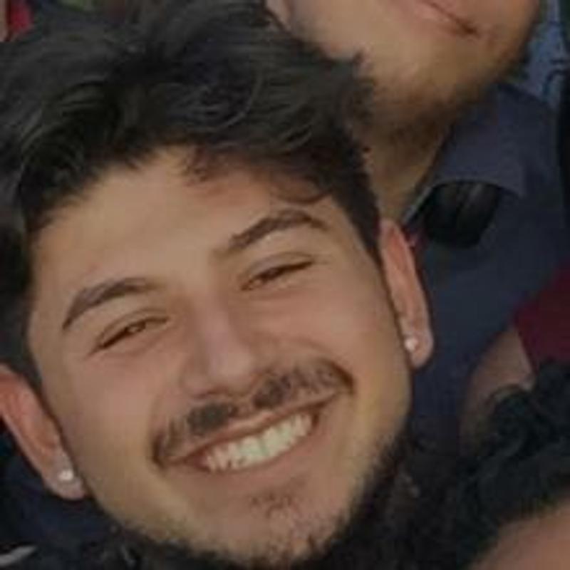 A smiling young person with curly hair, a mustache, a short beard, and earrings