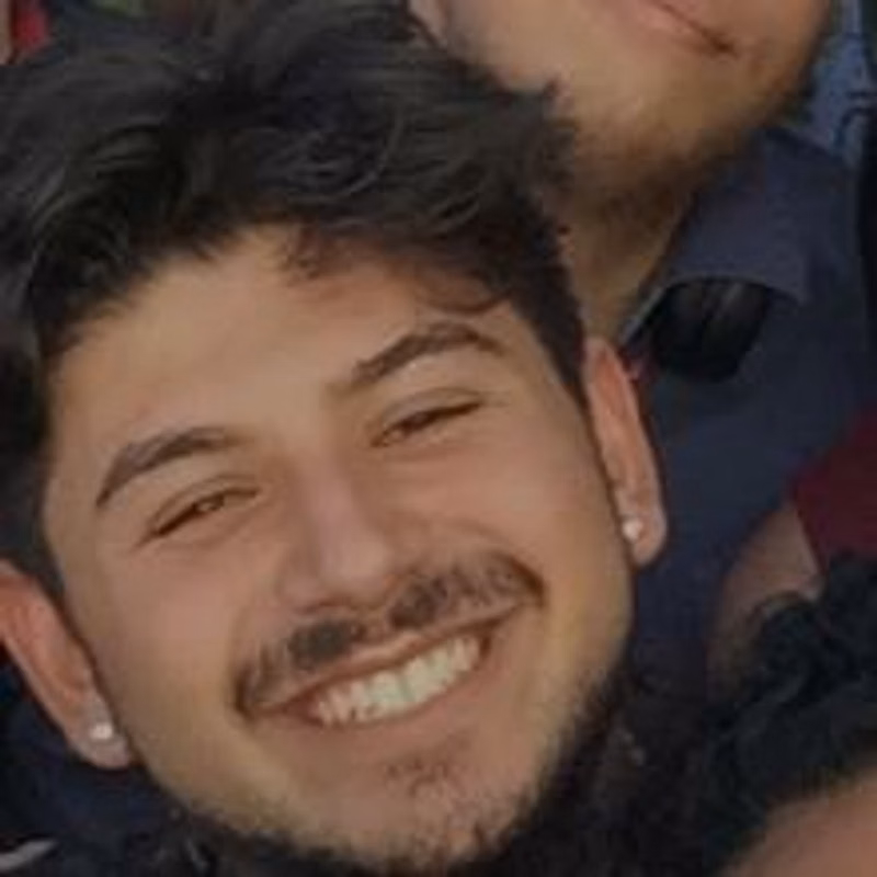 A smiling young person with curly hair, a mustache, a short beard, and earrings