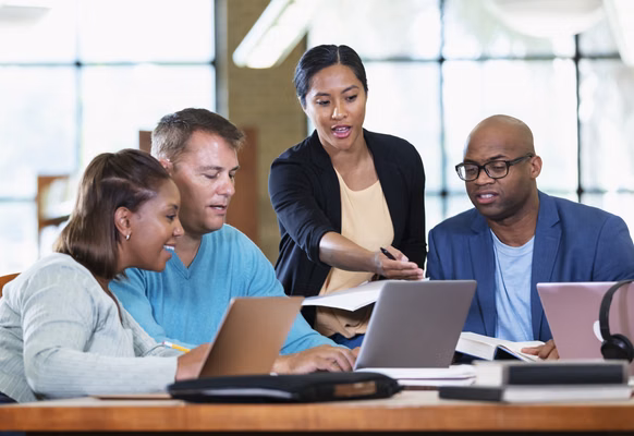 A multiracial group of four adults working together on a project at a table in the library. They are adult students or business people in a meeting, with books and laptops.